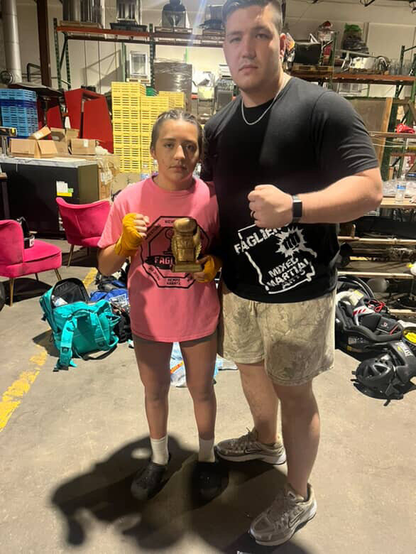 Young student in a pink Faglier's MMA shirt holding a trophy beside a coach